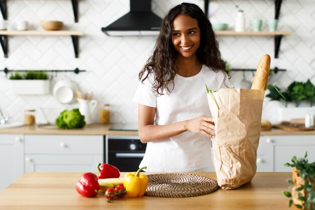 笑顔のムラートの女性は、モダンな白いキッチンでバゲットと野菜のパッケージを保持しています。