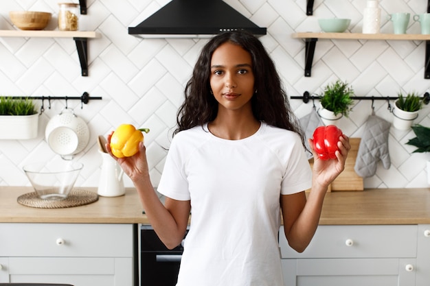 Smiled mulatto woman with loose hair is holding red and yellow peppers in hands near the kitchen desk on the modern white kitchen dressed in white t-shirt