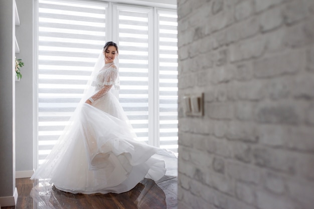 Улыбающаяся красавица оборачивается в комнате возле белой кирпичной стены, одетая в модное платье, свадебная мода