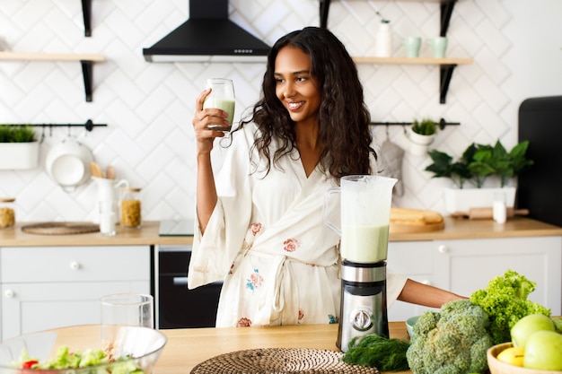 笑顔のかなりムラートの女性は、新鮮な野菜とテーブルの近くに緑のスムージーを保持している白いモダンなキッチンにルーズな髪のナイトウェアを着て、ガラス製品を探して