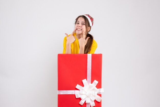 smiled young girl with santa hat standing behind big xmas gift on white