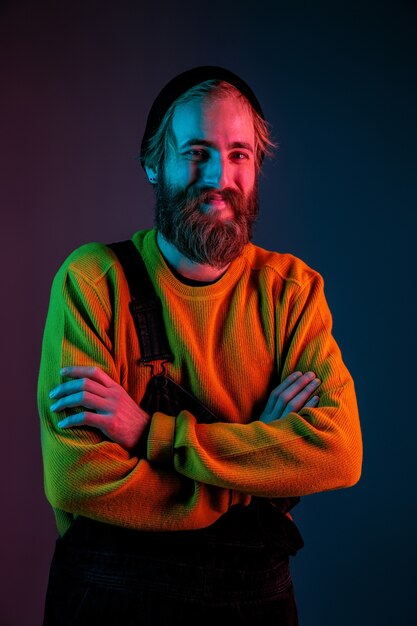 Smiles, hands crossed. Caucasian man's portrait on gradient studio background in neon light. Beautiful male model with hipster style. Concept of human emotions, facial expression, sales, ad.