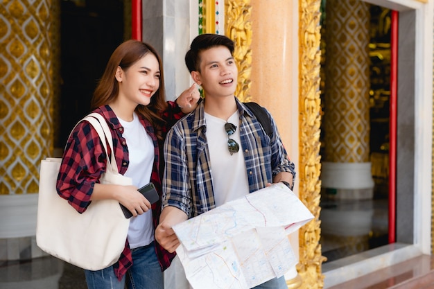 Smiley Asian couple tourist backpackers standing in beautiful Thai temple, pretty woman holding paper map and handsome man check in smartphone with happy on vacations