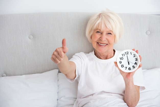 Smiley elder woman holding a clock in her bed