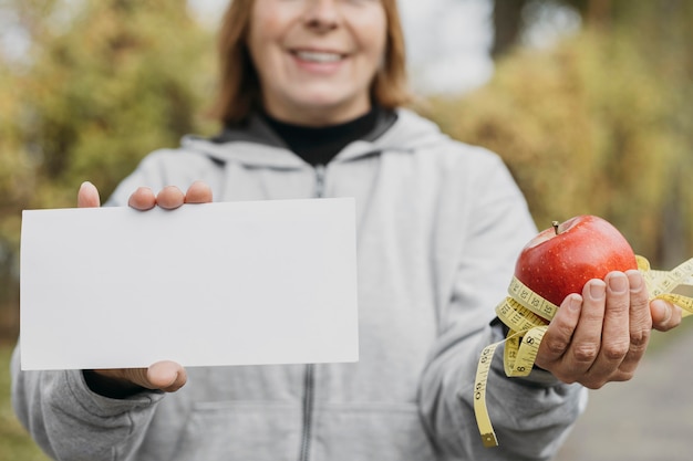 Smiley elderly woman holding apple and paper outdoors while working out