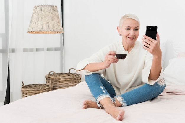 Smiley elderly woman holding cup of coffee and looking at phone in bed