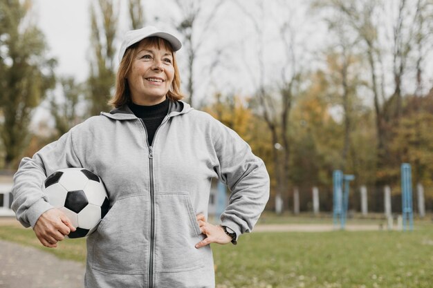 Смайлик пожилая женщина держит футбол на открытом воздухе во время тренировки