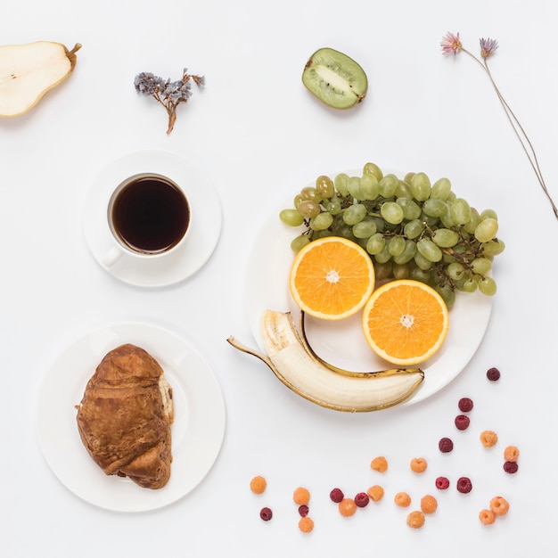 白い皿の上の果物とコーヒーで作られたスマイリーフェイス。クロワッサンとコーヒーの白い背景で隔離