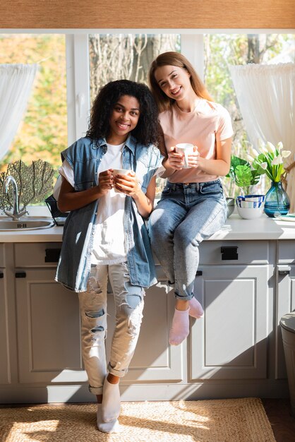 Smiley female friends posing in the kitchen while holding mugs