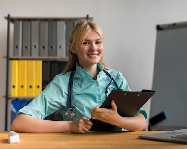 Free photo smiley female nurse in the office with notepad and stethoscope