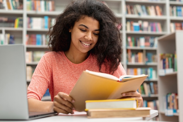 Улыбающаяся девушка в библиотеке изучает и использует ноутбук