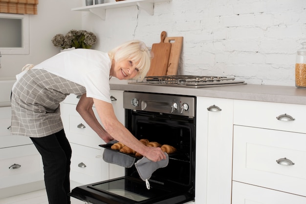 Улыбающаяся пожилая женщина держит поднос с круассанами в духовке