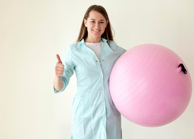 Смайлик физиотерапевт держит мяч для упражнений и показывает палец вверх