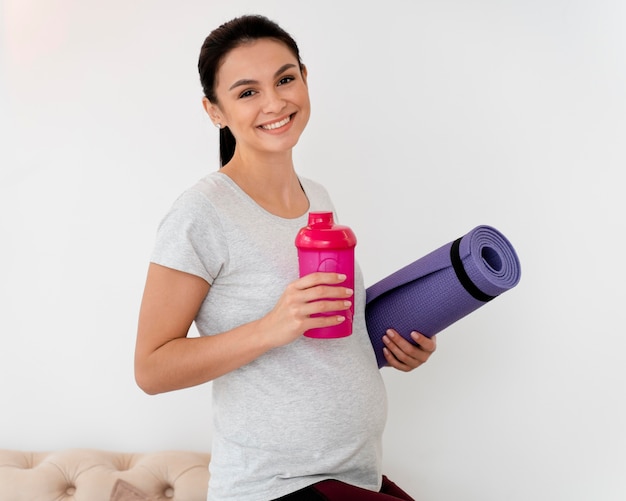 Улыбающаяся беременная женщина, держащая фитнес-коврик и бутылку воды