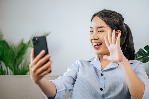 Smiley pretty teenage sitting on sofa and use smartphone to video call or selfie,  during quarantine covid-19 self isolation at home