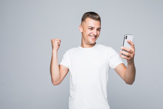 Smiley strong young man is wearing a white t-shirt and taking a selfie with a silver smartphone.