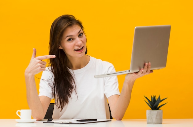 Smiley woman at desk holding up and pointing at laptop