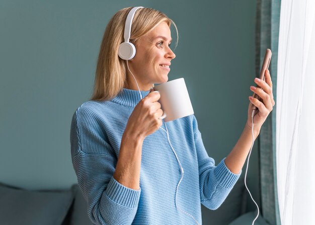 Smiley woman using smartphone and headphones at home while having coffee during the pandemic
