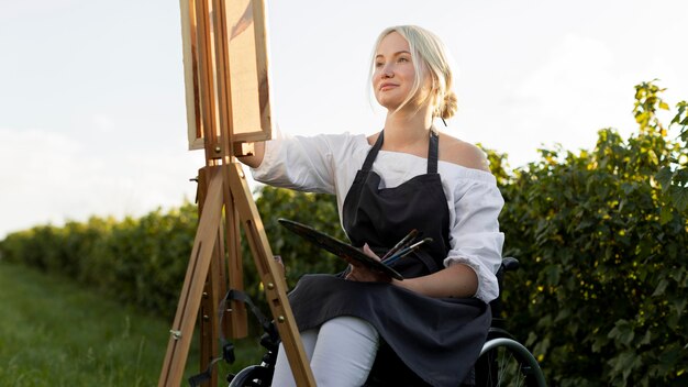 Smiley woman in wheelchair outdoors in nature with canvas and palette