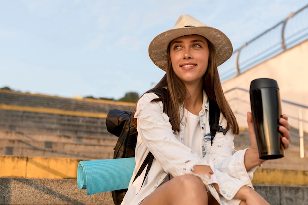 Smiley woman with backpack and hat holding thermos while traveling