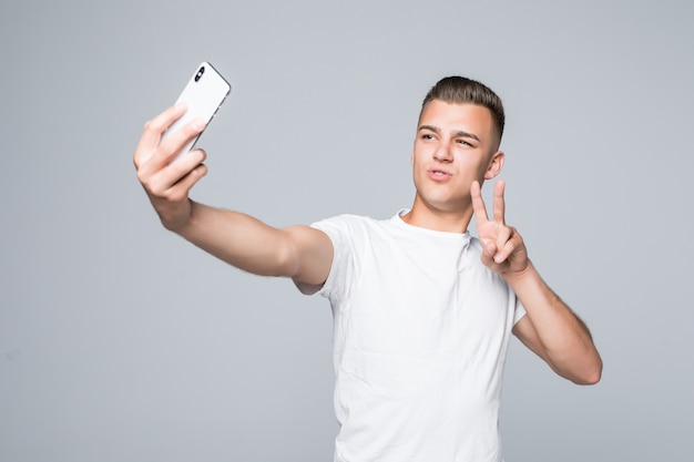 スマイリーの若い男は白いTシャツを着て、銀色のスマートフォンで自分撮りの勝利のサインを取っています。