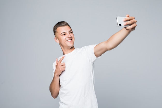 スマイリーの若い男は白いTシャツを着て、銀色のスマートフォンで自分撮りをしています。