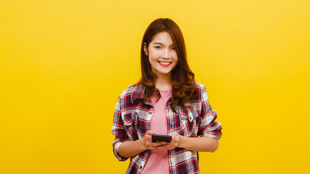 Smiling adorable Asian female using phone with positive expression, smiles broadly, dressed in casual clothing and looking at camera over yellow wall. Happy adorable glad woman rejoices success.