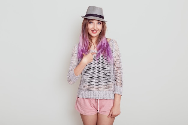 Smiling adorable female in hat and pointing at herself with index finger, wearing shirt and stylish hat, being amazing that choose her, isolated over white wall.