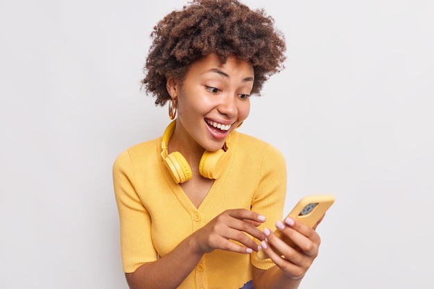 스마트폰을 통해 온라인으로 곱슬머리를 하고 웃고 있는 사랑스러운 10대 소녀는 현대 기술에 중독된 응용 프로그램을 사용하여 목 캐주얼 노란색 점퍼 흰색 벽에 스테레오 헤드폰을 착용합니다.