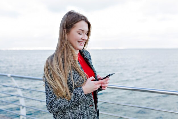 Smiling adorable woman in grey coat and red shirt scrolling smartphone on the embankment and enjoys good weather