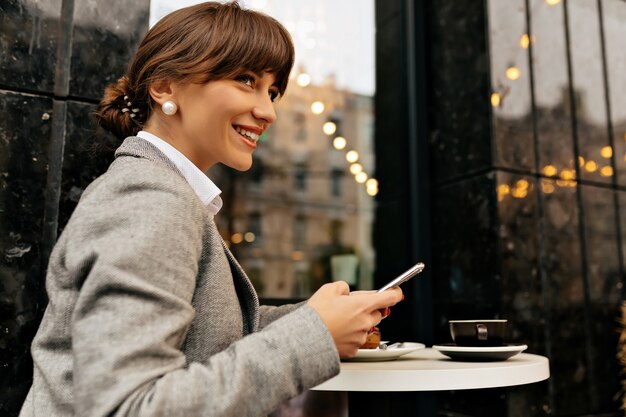 Smiling adorable woman wearing grey jacket sitting in open air cafe is using smartphone and waiting for meeting on background of city lights High quality photo