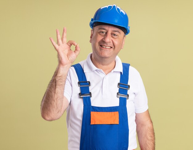 Smiling adult builder man in uniform gestures ok hand sign isolated on olive green wall