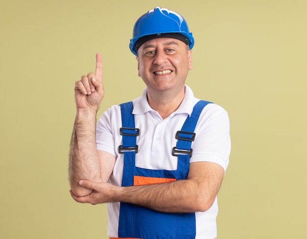 Smiling adult builder man in uniform points up isolated on olive green wall