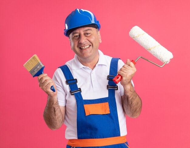 Smiling adult caucasian builder man in uniform holds paint brush and roller brush on pink