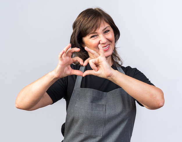 Smiling adult caucasian female barber in uniform gesturing heart sign isolated on white background with copy space