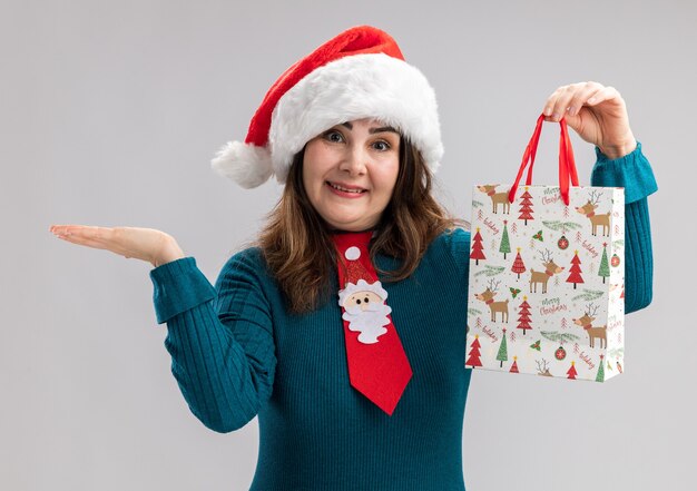Smiling adult caucasian woman with santa hat and santa tie holding paper gift box and keeping hand open isolated on white background with copy space