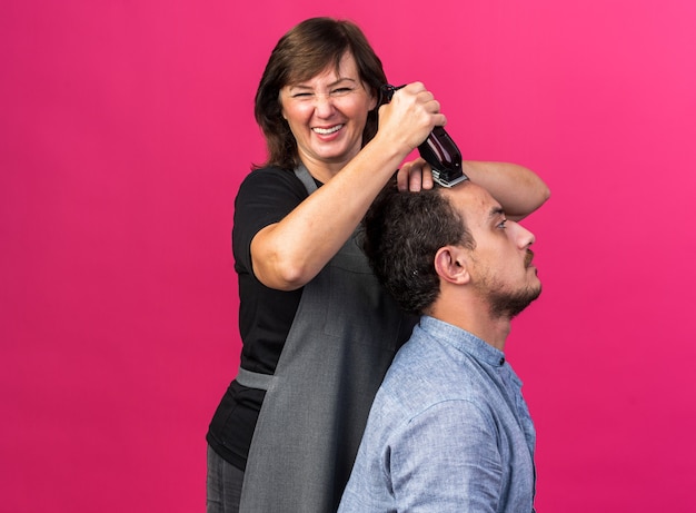 smiling adult female barber in uniform doing haircut for young man with hair clipper isolated on pink wall with copy space