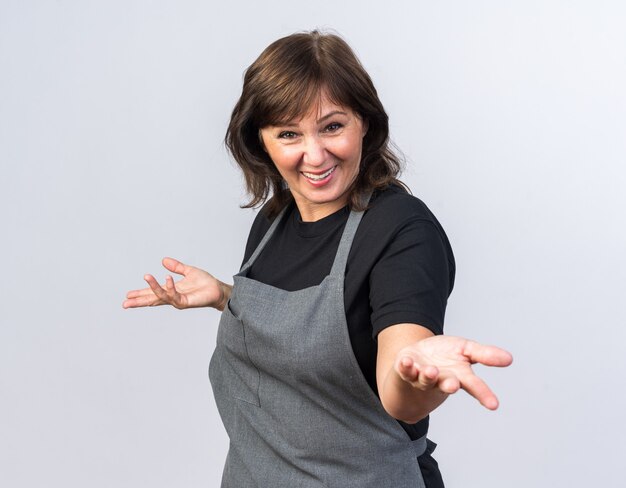 smiling adult female barber in uniform holding hands open and looking at front isolated on white wall with copy space