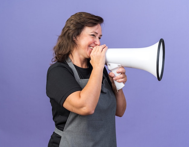 Free photo smiling adult female barber in uniform holding loud speaker and looking at side isolated on purple wall with copy space