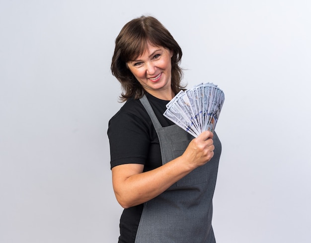 smiling adult female barber in uniform holding money isolated on white wall with copy space