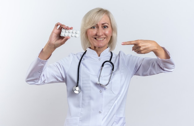 smiling adult female doctor in medical robe with stethoscope holding and pointing at medicine tablet in blister pack isolated on white wall with copy space