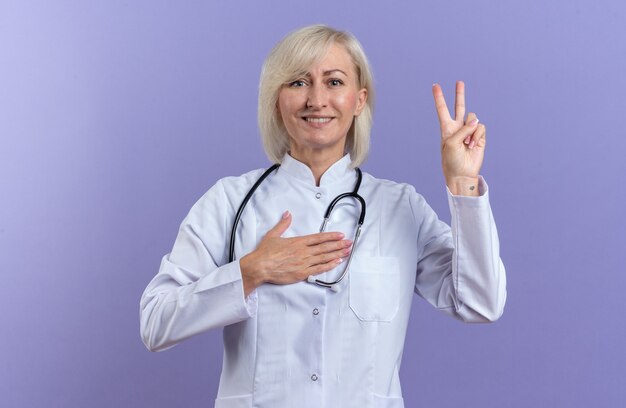 smiling adult female doctor in medical robe with stethoscope putting hand on chest and gesturing victory sign isolated on purple wall with copy space