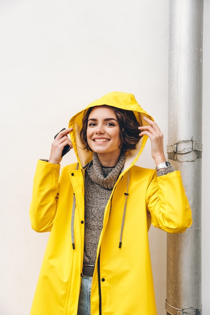 Smiling adult girl in yellow coat putting hood on head posing on camera with candid smile being happy