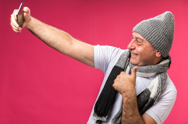 복사 공간이 분홍색 벽에 고립 된 selfie를 복용 전화를보고 겨울 모자 엄지 손가락을 입고 목에 스카프와 성인 아픈 백인 남자를 웃고