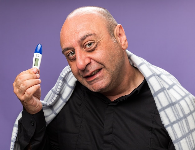 smiling adult ill caucasian man wrapped in plaid holding thermometer isolated on purple wall with copy space