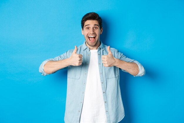 Smiling adult man showing thumbs up with excited face, like something awesome, approving product, standing against blue background.