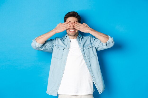 Smiling adult man waiting for surprise, covering eyes with hands and anticipating, standing against blue background in casual clothes