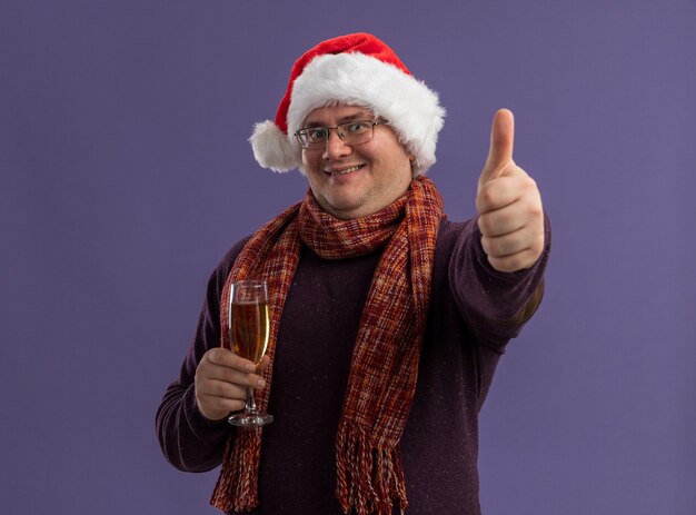 Smiling adult man wearing glasses and santa hat with scarf around neck holding glass of champagne  showing thumb up isolated on purple wall