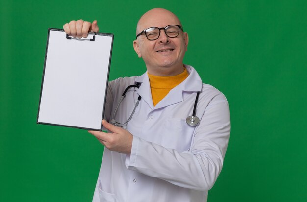 Smiling adult man with glasses in doctor uniform with stethoscope holding clipboard and looking 
