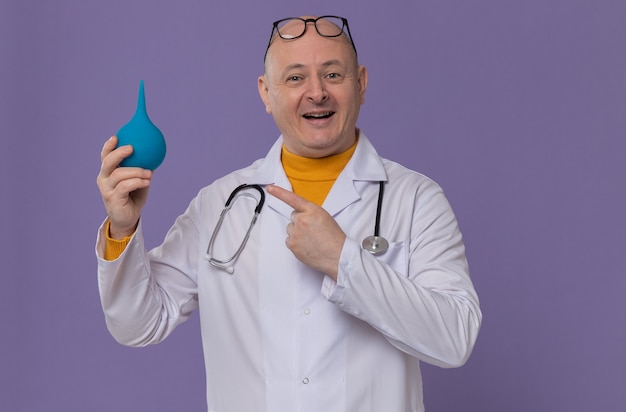 Foto gratuita uomo adulto sorridente con gli occhiali in uniforme da medico con lo stetoscopio che tiene e guarda il clistere